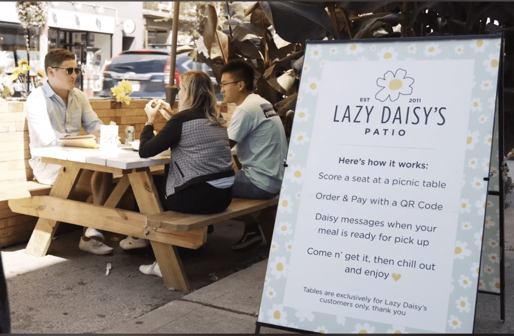 A Frame sign in front of Lazy Daisy's Cafe patio in Toronto with instructions on how to Scan the QR code to view the retaurant menu, then place an order from your mobile phone and pay. When your order is ready, you will get a text to pick it up